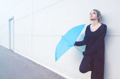 Thoughtful young woman with umbrella standing against wall