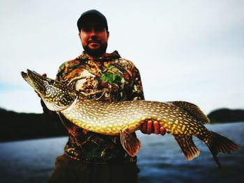 Fisherman holding pike by sea against sky