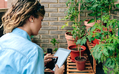 Woman touching digital tablet while caring plants of urban garden on terrace