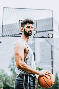 Side view of confident young bearded male player with tattooed arm holding basketball ball and looking away while standing on street playground
