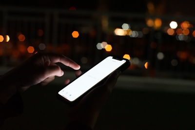 Cropped hand holding using phone against defocused lights at night