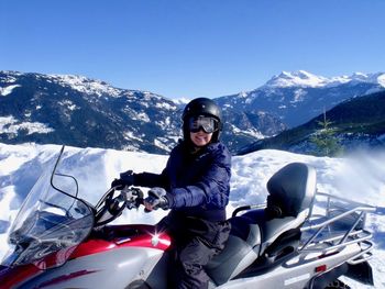 Portrait of smiling woman sitting on snowmobile at mountains against blue sky
