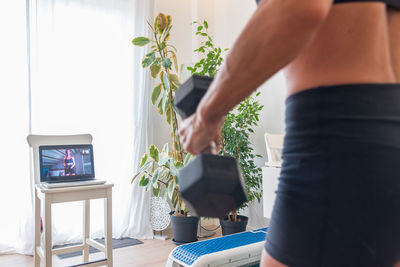 Back view of unrecognizable female athlete watching online video on laptop and doing exercises with dumbbells during training at home