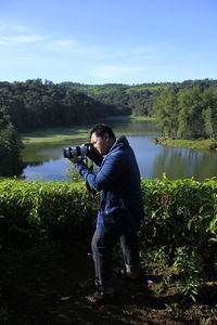 Man photographing while standing by lake against trees