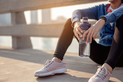 Low section woman holding water bottle while sitting on footpath