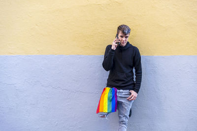 Non binary person with an lgbt rainbow flag talking on the phone outdoors.