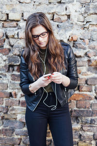 Young woman using phone while standing against brick wall