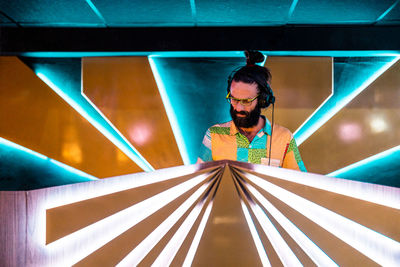 Concentrated young bearded male dj in eyeglasses and headphones standing at illuminated counter in modern nightclub