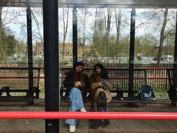 People sitting by railing against trees seen through glass window