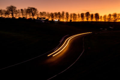 Illuminated road by silhouette trees against sky at sunset