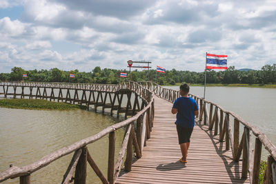 Rear view of boy walking on footbridge against sky