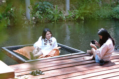 Woman photographing friend sitting on lake in forest