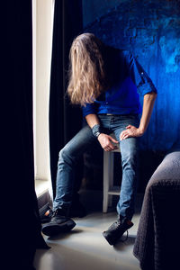 Man with long hair and beard sits at home on a chair by the window in a blue shirt and jeans