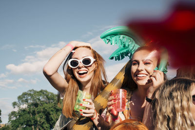 Cheerful female friends enjoying drinks at music concert against sky in summer