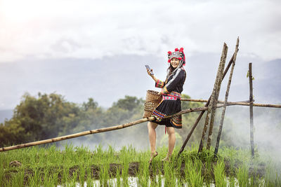 Portrait of smiling woman holding mobile phone and basket while standing on agricultural field
