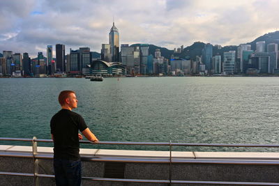 Rear view of man standing by railing against river with cityscape in background