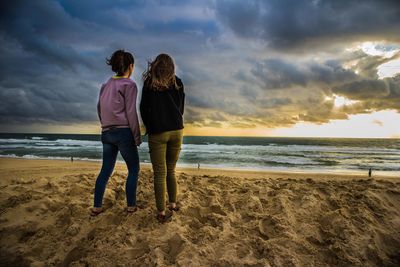 Rear view of friends standing on beach against sky during sunset