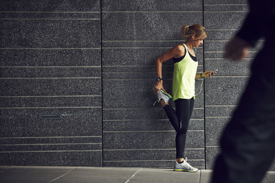 Young jogger stretching with cell phone against grey wall