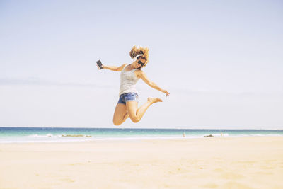 Smiling woman listening music while jumping at beach during summer
