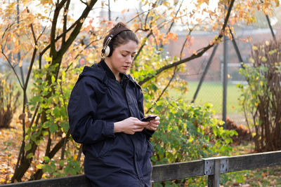 Young woman listening music while standing against trees in park