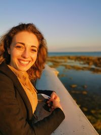 Portrait of smiling young woman using mobile phone at beach against sky