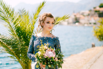 Bride holding bouquet while standing against sea