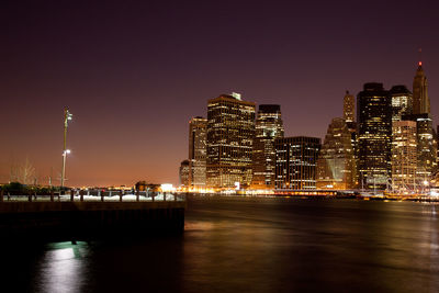 Illuminated buildings by river against clear sky at night