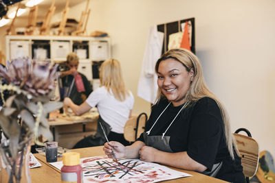 Portrait of smiling young female student painting on canvas while sitting at table in art class