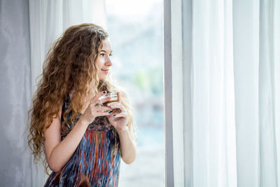 Woman having tea while looking through window while standing at home