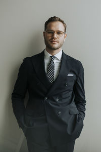 Portrait of confident young businessman standing with hands in pockets against white wall