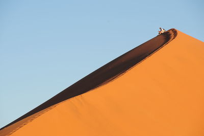 Man sitting on sand dunes at desert against clear sky