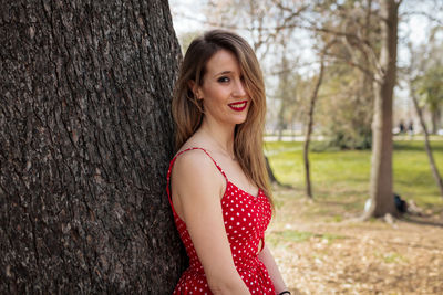 Portrait of smiling woman standing against tree trunk in park