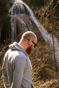 Man wearing hooded shirt standing against waterfall at forest