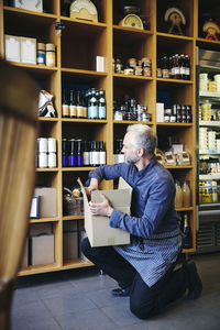 Mature salesman unpacking box and arranging beauty products on rack in deli