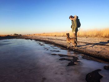 Man looking at dog while standing by wet field against clear sky