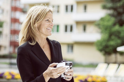 Happy businesswoman looking away while holding smart phone and disposable coffee cup outdoors