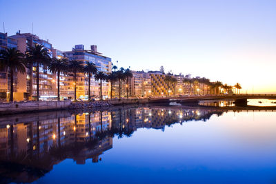 Illuminated buildings by lake against sky in city at dusk