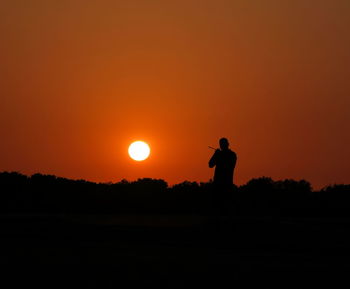 Silhouette man photographing on field against orange sky