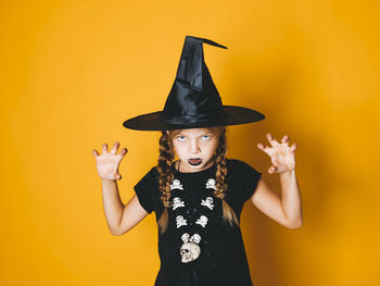 Portrait of girl wearing witch hat gesturing against yellow background during halloween