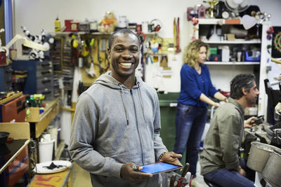 Portrait of smiling mid adult volunteer holding digital tablet with colleagues working in background at workshop