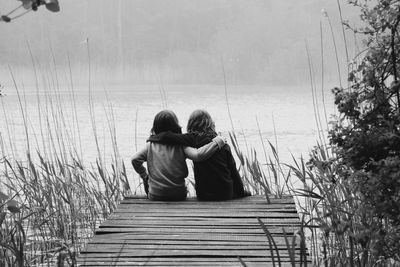 Rear view of friends with arm around sitting on pier by river during foggy weather