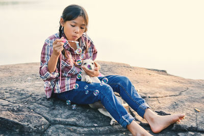 Girl blowing bubbles while sitting with puppy on rock against lake
