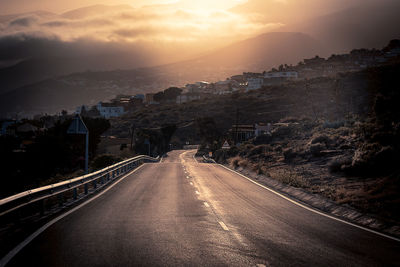 Road amidst buildings in city against sky during sunset