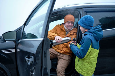 Happy father and son family are resting on side of road on road trip drinks hot tea from thermos