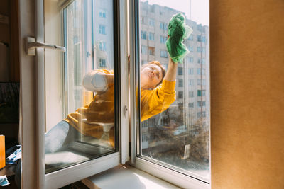 Caucasian woman of fifty in a yellow sweater and jeans washes a dusty window in apartment. 