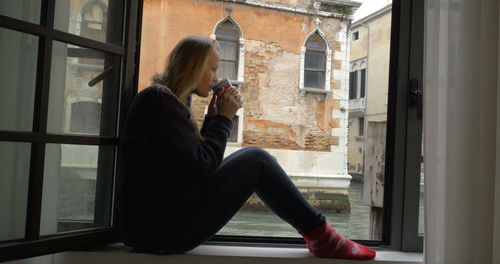 Woman photographing through smart phone while sitting on window