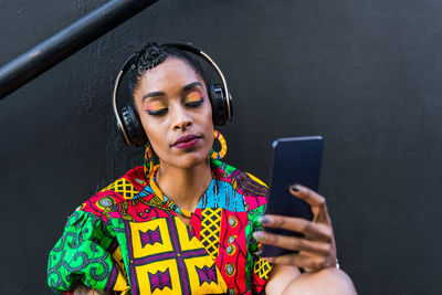 Young ethnic female in colorful stylish outfit listening to music in wireless headphones and browsing social media on cellphone
