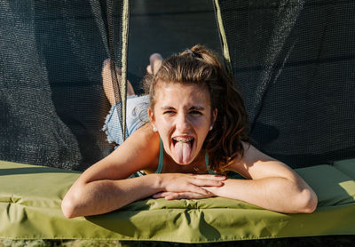 Horizontal portrait of a curly brunette teenager lying on a trampoline showing her tongue to the camera and coming through the net. the girl wears a green top and looks tired after jumping.
