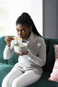 Young woman eating noodles while sitting on sofa at home