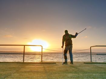 Man hiker with backpack stand alone and watching romantic colorful sunrise above wooden sea bridge